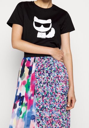 Person iført en sort t-shirt med hvid kat med solbriller grafisk print og flerfarvet plisseret nederdel med abstrakte mønstre.