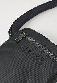 Borsa a tracolla nera in nylon con chiusura a zip e logo "BOSS" in rilievo. La tracolla è texturizzata per una migliore presa.