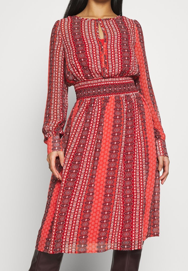 Femme portant une robe rouge à motifs, longueur genou, à manches longues, avec une encolure goutte d'eau et une taille cintrée, debout devant un fond uni.