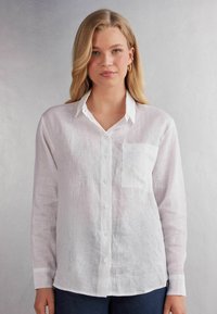 Bílá lněná košile s dlouhým rukávem, zapínáním na knoflíky vpředu, jednou náprsní kapsou a volným střihem. Hladká textura a jemné, přírodní tkaní.