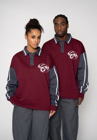 Bordeauxrote und graue Langarm-Poloshirts mit gestreiften Akzenten, Knöpfen am Kragen und einem Logo auf der Vorderseite. Locker sitzend, kombiniert mit grauen Hosen.