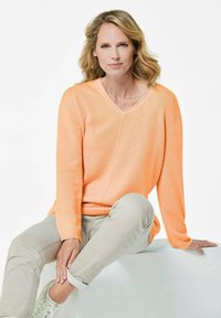 Pfirsichfarbener V-Ausschnitt-Pullover aus weichem Strick, mit langen Ärmeln und entspannter Passform, kombiniert mit hellbeigen Hosen und weißen Sneakers.