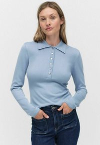 Kvinna med blont hår som bär en ljusblå långärmad kragtröja med knappar och mörkblå jeans, händerna i framfickorna.