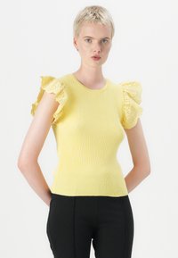 Gul ribbestrikket topp med korte, dekorative blondermerker. Tettsittende design, rund hals, kombinert med svarte bukser.