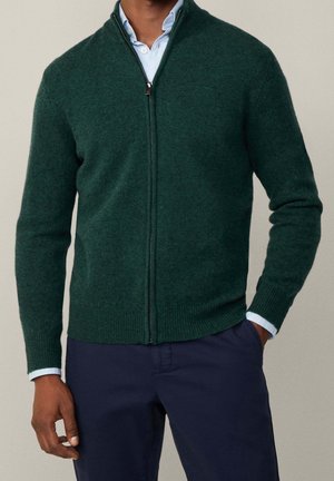 Homme portant un pull zippé vert foncé par-dessus une chemise bleu clair à col, et un pantalon bleu marine, debout avec une main dans la poche.