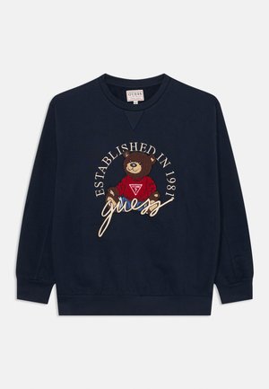Marineblå hettegenser med ribbede mansjetter, med en brodert bjørn i rød skjorte, omgitt av teksten "ETABLERINGSÅR 1981" og "guess".