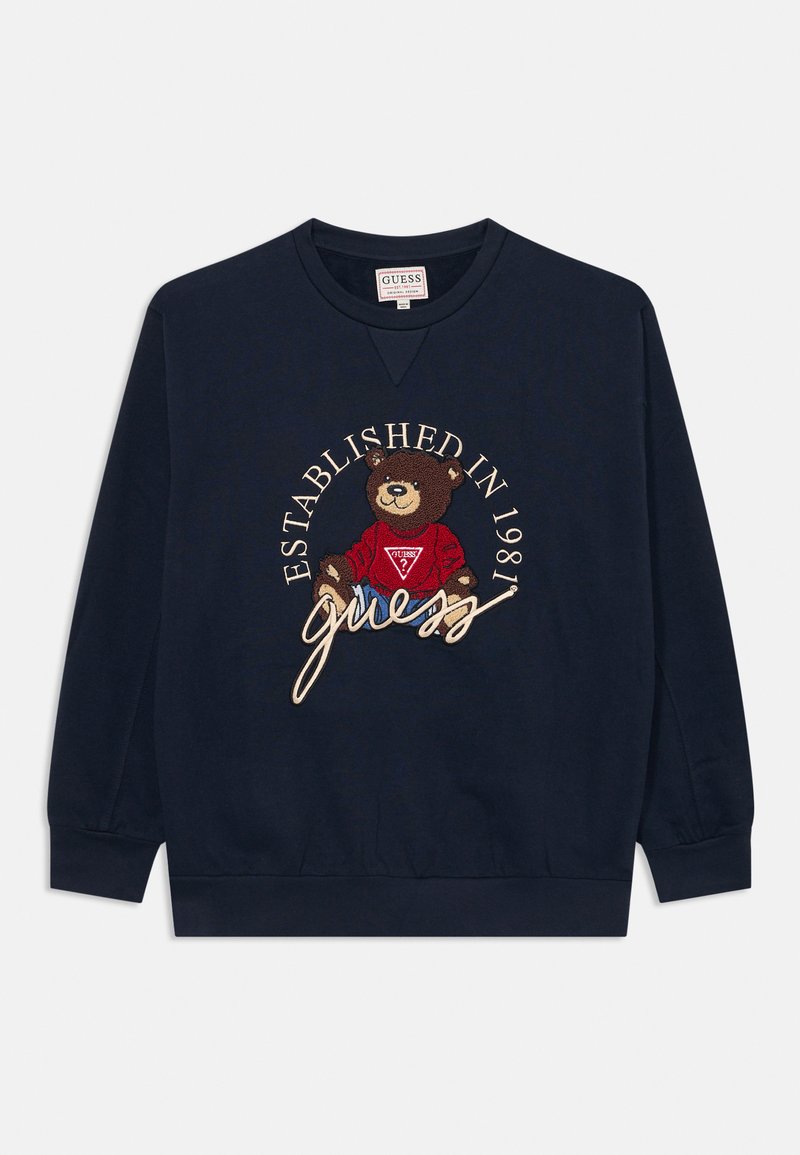 Sweatshirt marine avec des poignets côtelés, présentant un ours brodé portant une chemise rouge, entouré du texte "ÉTABLI EN 1981" et "guess".