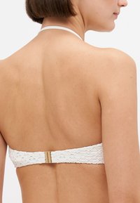 Bela bikini zgornji del ima teksturiran material s pikčasto obliko, halterski dizajn in zlat sponko na hrbtu.