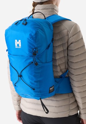 Personne portant une doudoune beige et un sac à dos de randonnée Millet bleu vif avec des bretelles réglables et un système de cordons externe.