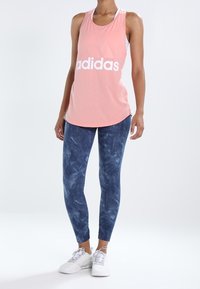 Canotta senza maniche rosa chiaro con logo bianco "adidas", abbinata a leggings texturizzati blu navy e scarpe da ginnastica bianche.