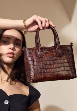 Braune, krokodilgeprägte Handtasche mit zwei Griffen, goldener Markenprägung, strukturiertem Design und glatter Textur; von einer Person vor einem neutralen Hintergrund gehalten.