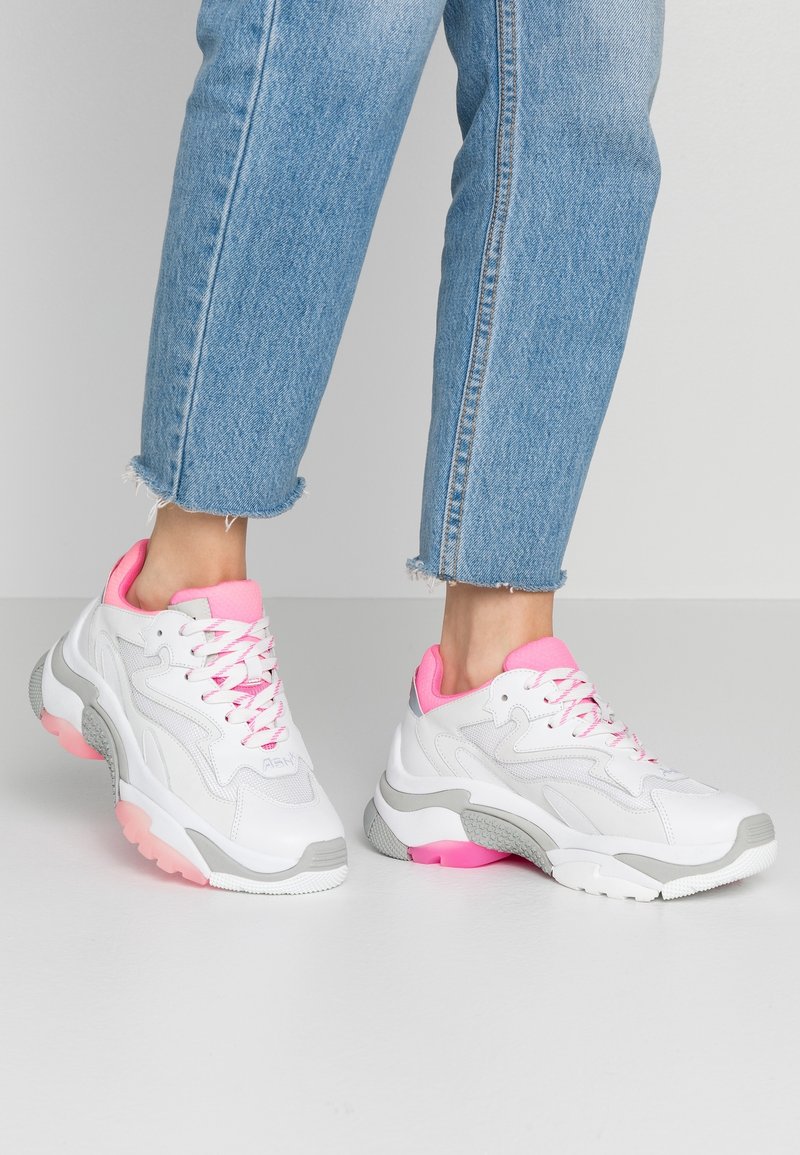 Chaussures de sport blanches avec des accents roses et des détails gris. Composées de mesh et de matériau synthétique, avec une semelle épaisse et des lacets contrastants.