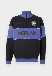 Sort og lilla halv-zip sweatshirts med "REPLAY"-logo på brystet og Replay FC-crest med tre stjerner øverst til venstre.
