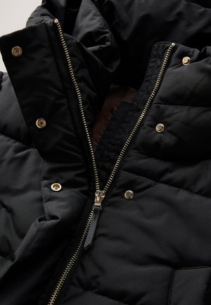 Sort quiltet jakke med metal lynlås delvist åben og fire synlige guld trykknapper på polstret stof.