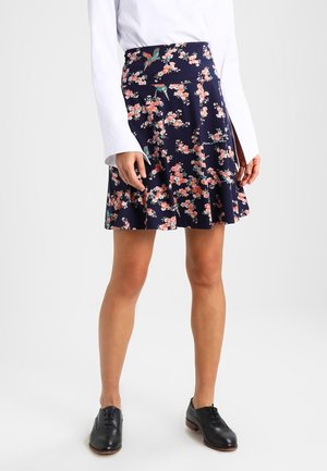 Femme portant une jupe fleurie bleu marine associée à une chemise blanche à manches longues et des chaussures à lacets noires, debout devant un fond uni.