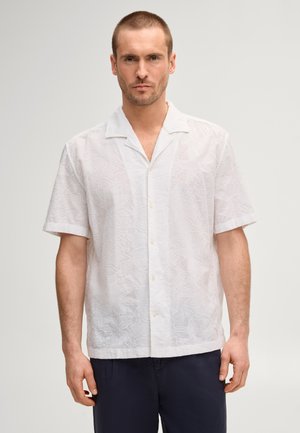 Mann trägt ein weißes, kurzärmeliges Hemd mit feiner Blumenstickerei und dunkler Hose, steht vor einem einfarbigen Hintergrund.