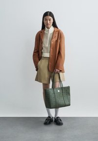 Borsa a tracolla verde con motivo in rilievo, abbinata a pantaloni beige e una giacca marrone strutturata sopra un maglione color crema. Mocassini neri e calze bianche.