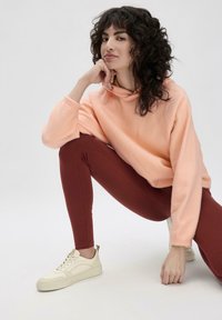 Meliert peachfarbenes Fleece-Sweatshirt mit Kapuze und Kordelzug, kombiniert mit bordeauxfarbenen, hoch taillierten Leggings und cremfarbenen Sneakers mit leichten Akzenten.