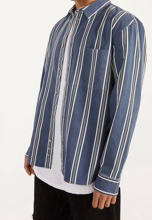 Mann trägt ein blaues Hemd mit weißen vertikalen Streifen über einem weißen T-Shirt und schwarzen Hosen, sichtbar von den Schultern bis zur Hüfte.