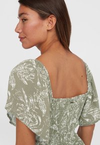 Robe verte à motifs avec de courtes manches bouffantes, dos carré, tissu texturé et motifs blancs complexes tout au long.