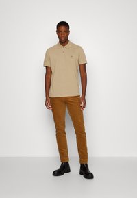 Polo bege com colarinho, mangas curtas; combinado com calças de veludo cotelê castanhas e botas pretas até ao tornozelo. Modelo em pé contra um fundo liso.