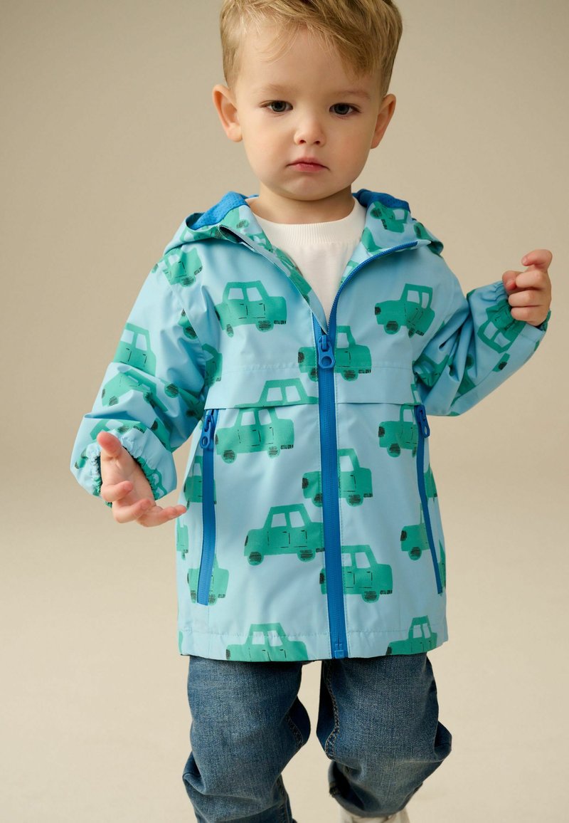 Young child wearing a blue jacket with car prints and jeans, standing with arms slightly raised against a plain background.