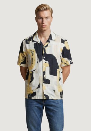 Hombre rubio con camisa de manga corta con botones y estampados abstractos en negro y beige, combinada con vaqueros azules, de pie con las manos detrás de la espalda.