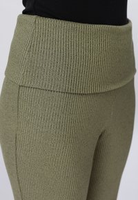 Κοντινό πλάνο σε ελαφρώς ριγωτό πλεκτό παντελόνι σε ελιά πράσινο χρώμα με φαρδιά, διπλωμένη μέση, φορεμένο με μαύρο τοπ.