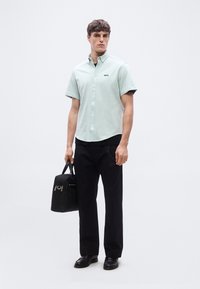 Άνδρας που φοράει ανοιχτοπράσινο πουκάμισο κοντού μανικιού με κουμπιά, μαύρο παντελόνι και παπούτσια, κρατώντας μια μαύρη τσάντα εργασίας σε ουδέτερο φόντο.