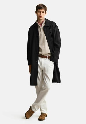 Joven de pie con las piernas cruzadas, vestido con abrigo negro, suéter beige con cremallera sobre camisa de cuadros roja, pantalones blancos, cinturón marrón y mocasines marrones.