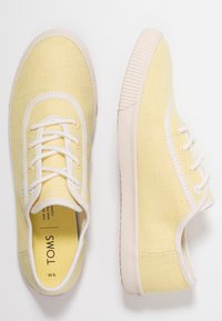 Baskets en toile jaunes avec des lacets blancs et une surface texturée. Présente une semelle en caoutchouc de couleur claire et un design à bout rond. Marque TOMS visible.
