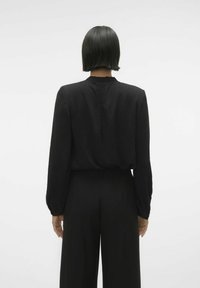 Blouse noire à manches longues, tissu texturé, avec un détail subtil de pli dans le dos, associée à un pantalon noir à larges jambes.