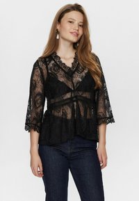 Blusa nera in pizzo con scollatura a V, motivo floreale e maniche a campana 3/4. Presenta un orlo a zig-zag e design a peplum. Indossata sopra jeans scuri.