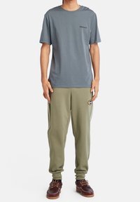 Šedé bavlněné tričko s logem, doplněné olivově zelenými tepláky s kulatou nášivkou a hnědými koženými botami se šněrováním.