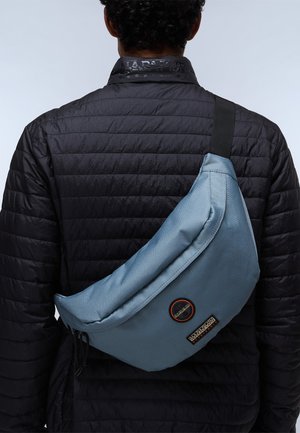 Borsa slouchy blu chiaro in tessuto, indossata a tracolla, con un patch circolare del logo e una tracolla nera, appoggiata su un giubbotto puffer nero.