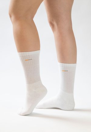 Benen met witte geribbelde sokken tot aan de kuit met kleine oranje en grijze strepen vlakbij de bovenkant, staand op een witte ondergrond tegen een effen achtergrond.