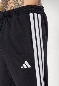 Black sweatpants with white three-stripe design, elastic waistband, and a small white logo. Soft texture, fitted around the waist.