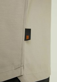 Bežinė marškinė su lygiu paviršiumi, žemutinėje siūlėje turi kontrastingą juodą ir oranžinį etiketę. Paprastas dizainas su tvarkingu siuvimo detalių apdailu.