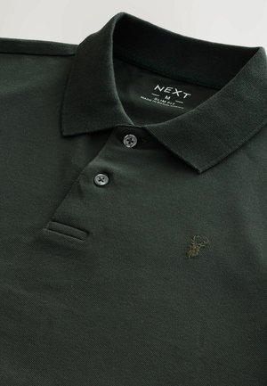 Camisa polo verde oscuro con cuello acanalado, tapeta de tres botones y un pequeño logotipo bordado de un ciervo en el lado izquierdo del pecho.