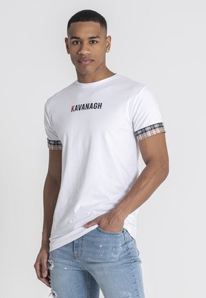 Camiseta de algodón blanca con el texto "KAVANAGH" en negro y rojo; mangas con un patrón de cuadros. Combinada con unos jeans rasgados de color azul claro.