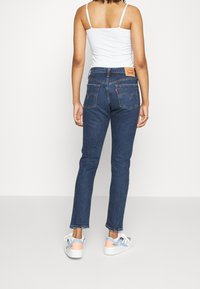 Jeans de mezclilla azules de tiro alto con corte recto. La parte trasera presenta la etiqueta del logo de Levi's y dos bolsillos traseros. Combinados con una camiseta sin mangas blanca y zapatillas.