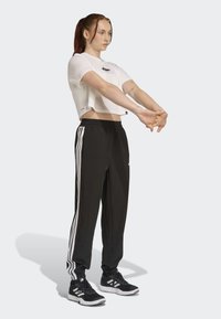 T-shirt blanc court avec logo, pantalon noir fuselé avec trois rayures blanches sur les côtés, et chaussures de sport noires avec des accents blancs.