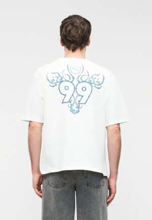 Bărbat cu spatele spre cameră, purtând un tricou alb cu un design de flacără albastră și numărul 99 pe spate, asortat cu blugi negri decolorați.