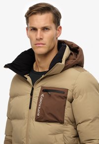 Vatteret jakke i beige med en brun brystlomme, sort foring og hætte. Har lynlås og logo på lommen. Glat tekstur.