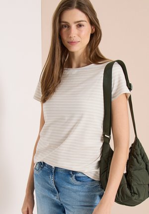 Ung kvinde med langt brunt hår iført en lyse stribet t-shirt og blå jeans, bærende på en mørkegrøn quiltet skuldertaske.