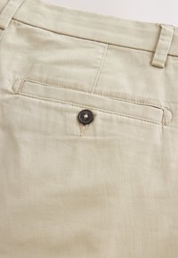 Beige fabric trousers showing the back pocket with a dark button and belt loop on the waistband.