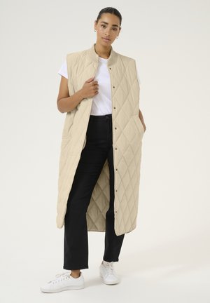 Gilet beige trapuntato senza maniche con colletto alto, dotato di bottoni a pressione e tasche laterali, indossato sopra una maglietta bianca e jeans neri.