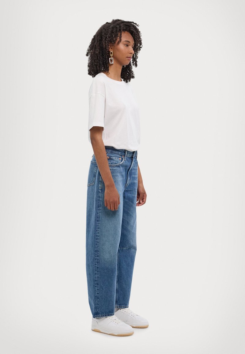 Weißes Kurzarmhemd kombiniert mit hoch taillierten blauen Jeans. Das Model trägt weiße Sneakers und große Creolen. Einfaches, lässiges Outfit.
