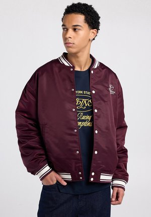 Jonge man kijkt naar links, draagt een donkerrode varsityjas met witte strepen en een marineblauw grafisch t-shirt, handen in de zakken.