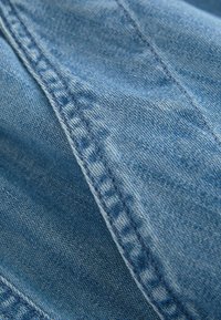 Denim-kangas, jossa on vaalea sininen sävy, näkyvillä ompeleilla ja tekstuurilla, kuvaa pehmeää, kestävää materiaalia ja klassista muotoilua.